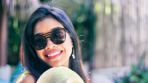 smiling woman wearing black sunglasses and holding a woven hat while standing outdoors