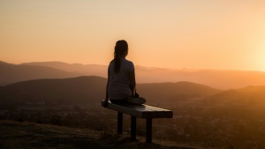 A woman sitting on a bench looking out at sunset vista.