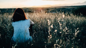 A young woman sitting in a field of grass and flowers watching the sunset.