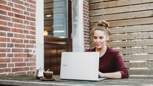 a woman seated at a wooden bench with a laptop and coffee with a brick wall and a door in the background