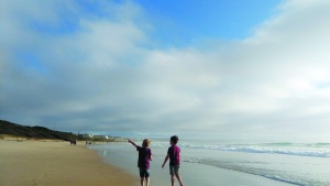 Two young boys walking along a beach, one pointing at something in the distance to the other. Wispy clouds cover part of the bright blue sky!