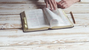 Photo of a marked up bible on a wooden table with a person's hands flipping through it. A pencil lays on the table next to the Bible.