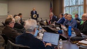 a group of men seated around a table watching a presentation by a man at a lectern