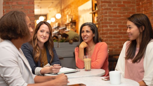 a group of four women dressed in business casual sitting at a table in a cafe with a brick wall that has a window looking into the rest of the cafe