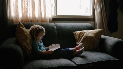 A little girls sitting on a couch looking at a book.