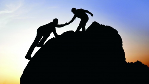 Silhouette of a person helping another person climb a boulder.