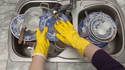 A person wearing yellow dish gloves and washing a sink full of dirty dishes.