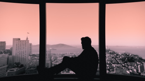A man sitting in a window sill looking out over a city skape.