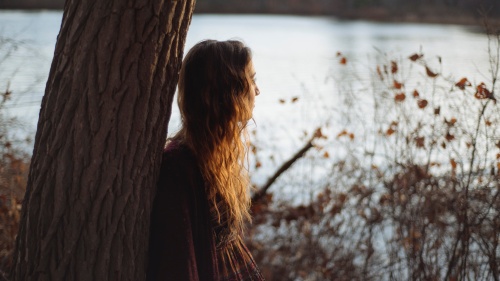 A young woman leaning against a tree looking out over a lake.