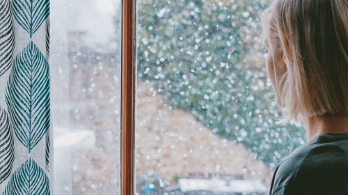 A young woman looking out a window as snow falls.