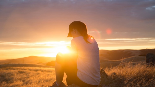 A woman praying while sitting on a rock with the sun setting in the background.