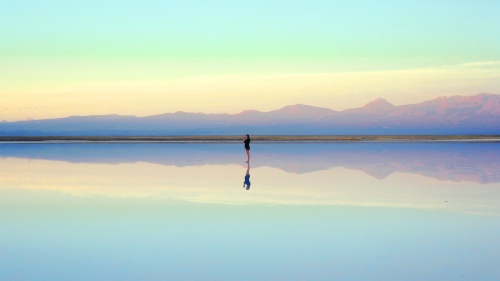 Photo of a person walking along the beach at low tide with their reflection in the water below.