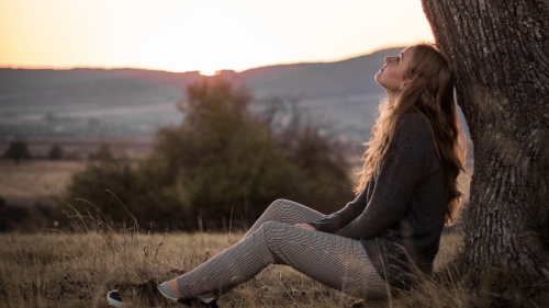 A young woman sitting on the ground leaning against a tree looking up toward the sky.