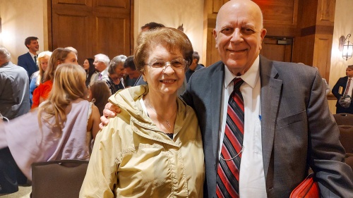 a couple stands in a meeting room with other people in the background and dressed in church wear