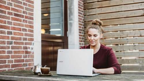 a woman seated at a wooden bench with a laptop and coffee with a brick wall and a door in the background