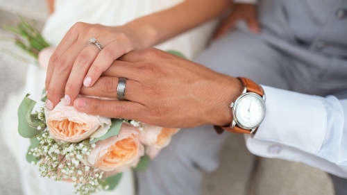a married couple's hands layered over a pink bouquet of flowers and wearing wedding rings