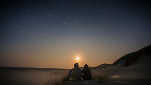 Two people sitting on a beach with the sun setting. 