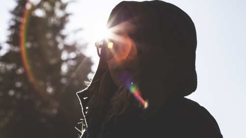 A girl wearing a jacket and hood looking at the camera with sunrays behind in head.