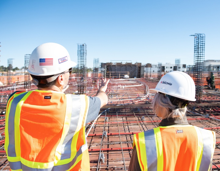 two people wearing construction hats and vests and looking over a construction site with their backs to the camera
