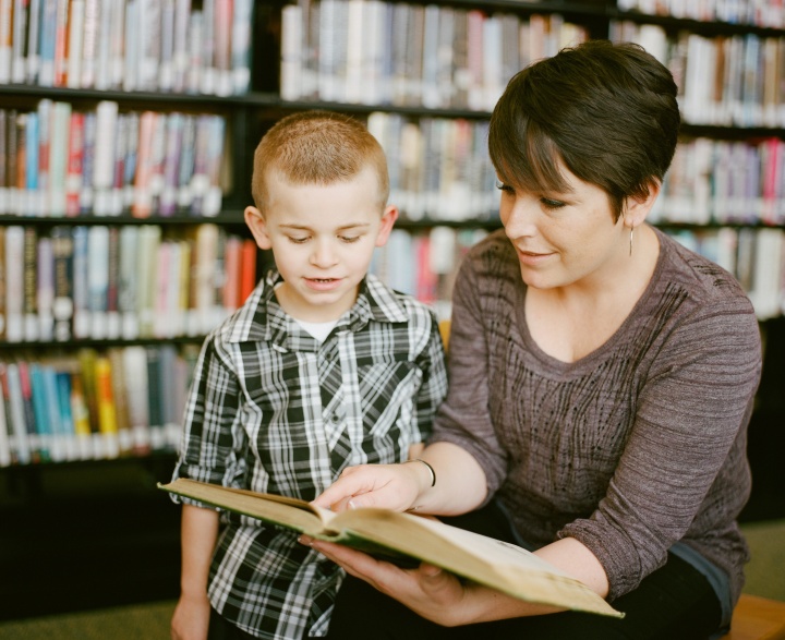 a woman and a child reading a book together with bookshelves in the background