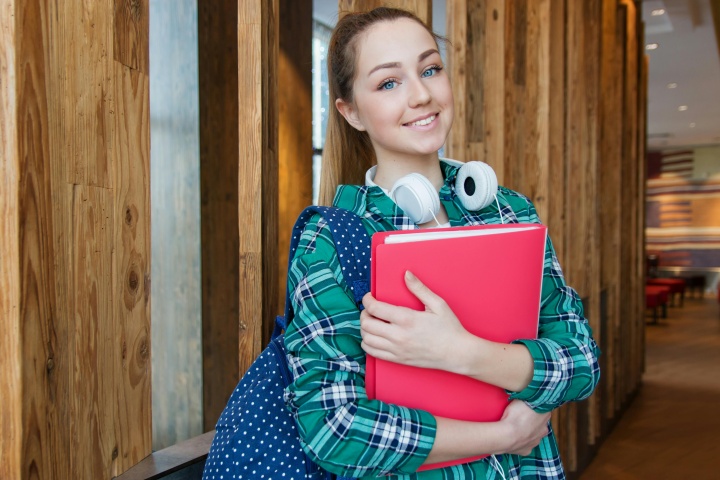 a woman with a backpack and arms full of school books standing in front of a wooden wall