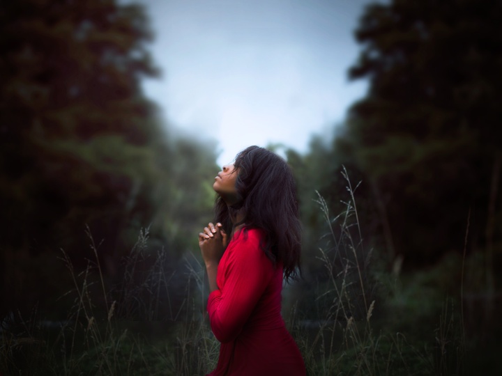 a woman wearing and red dress kneeling with folded hands outdoors amidst trees