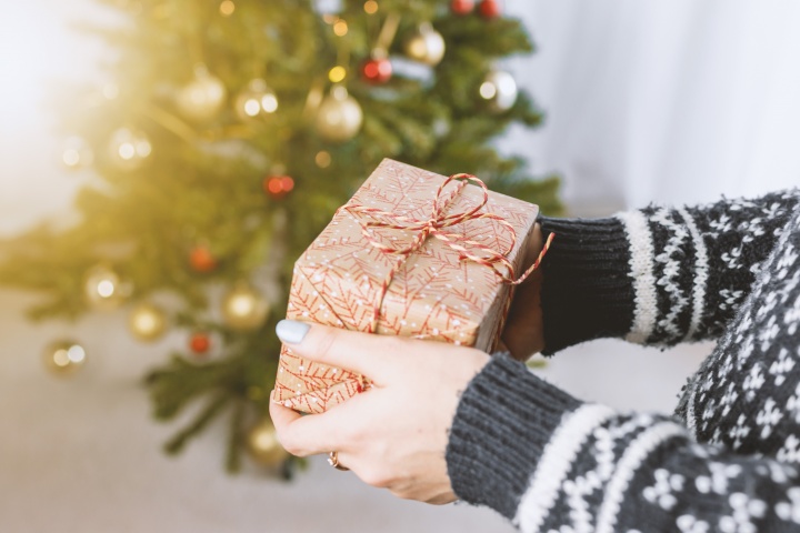 A person holding a wrapped gift in front of a Christmas tree.