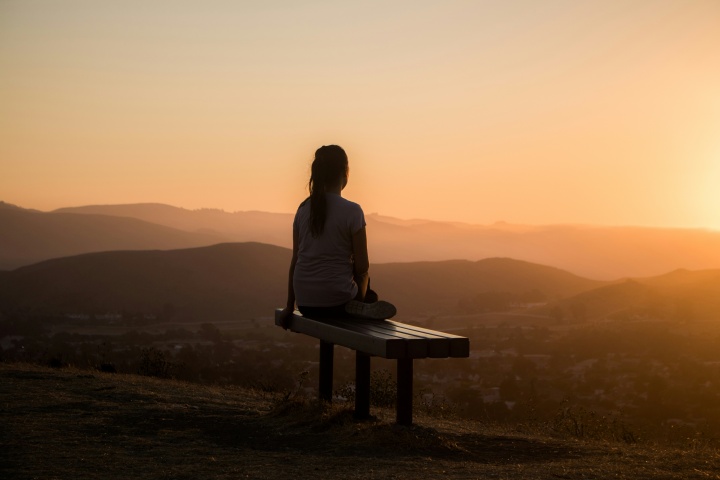 A woman sitting on a bench looking out at sunset vista.