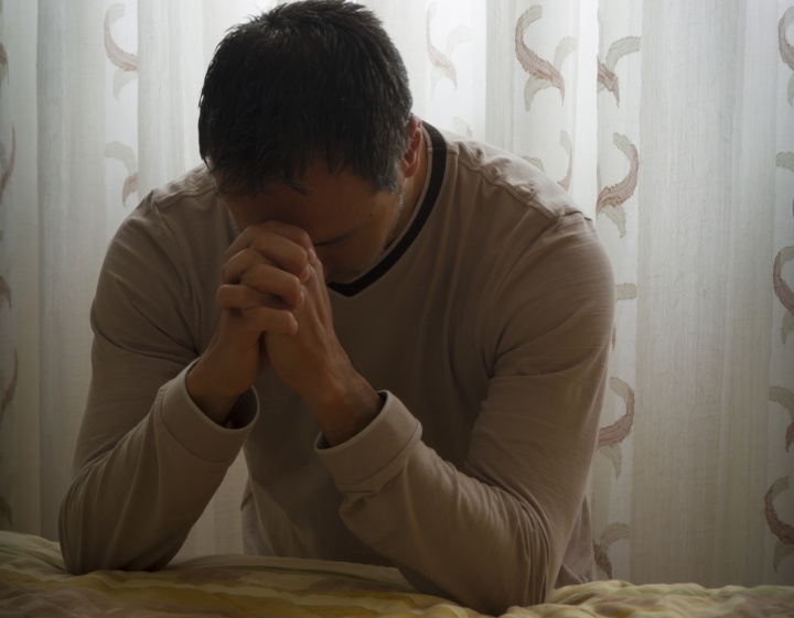 A man praying at the side of a bed.