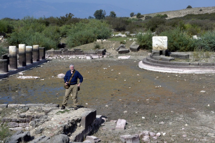 Scott Ashley standing at the edge of the harbor at Miletus, with the silted-in harbor and the remains of the monument behind him at the right. 