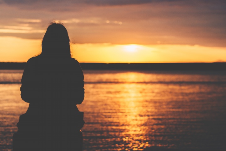 A woman sitting by a body of water as the sun is setting.