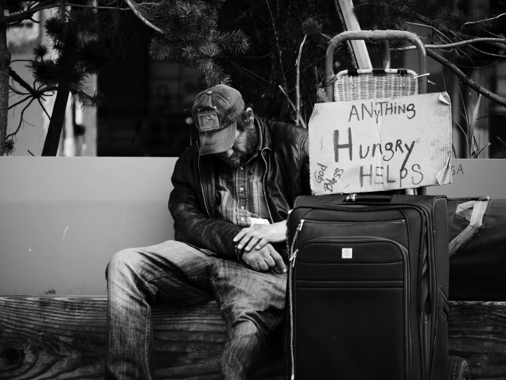 A homeless man sitting beside the road with a sign asking for help.