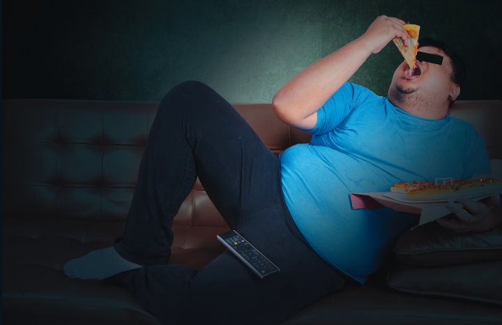 A lazy guy eating a slice of pizza on a coach while watching tv. 