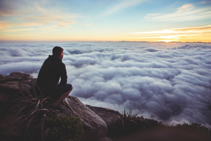 Photo of man thoughtfully squatting at the top of a cliff looking at the clouds.