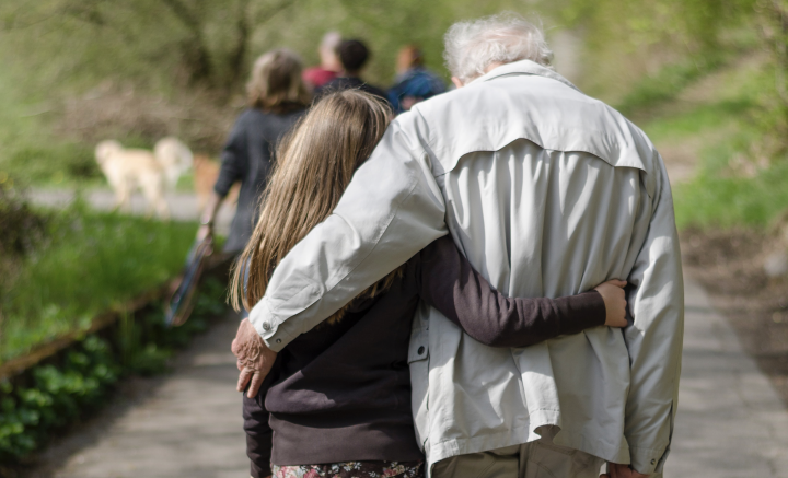 Photo of a young girl on the left and old man on the right, walking along a paved trail in a park, each with an arm around the other.