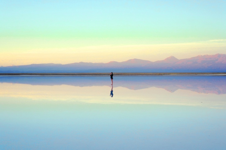 Photo of a person walking along the beach at low tide with their reflection in the water below.