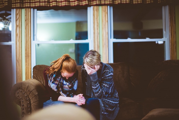 Photo of two women sitting on the couch grieving together. One woman has her hands clasped and one woman is leaning her face into her forhead.