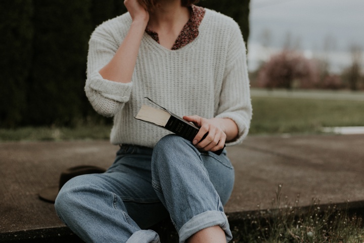 A girl in cropped mom jeans sits on the ground with a Bible in her lap. A wide brim hat sits next to her and she has a flowery collared shirt on under a white sweater.