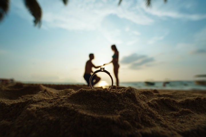 silhouette of man proposing to woman on a beach seen through a ring up close
