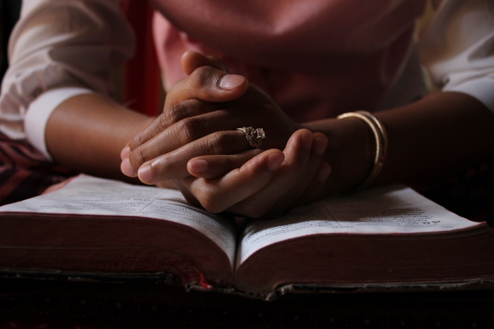 a pair of hands folded in prayer sitting atop an open Bible