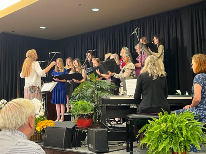 a choir standing on stage with a director standing in front of them and two ladies seated at a piano