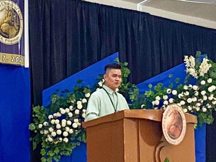 a man standing at a lectern with a blue and floral backdrop