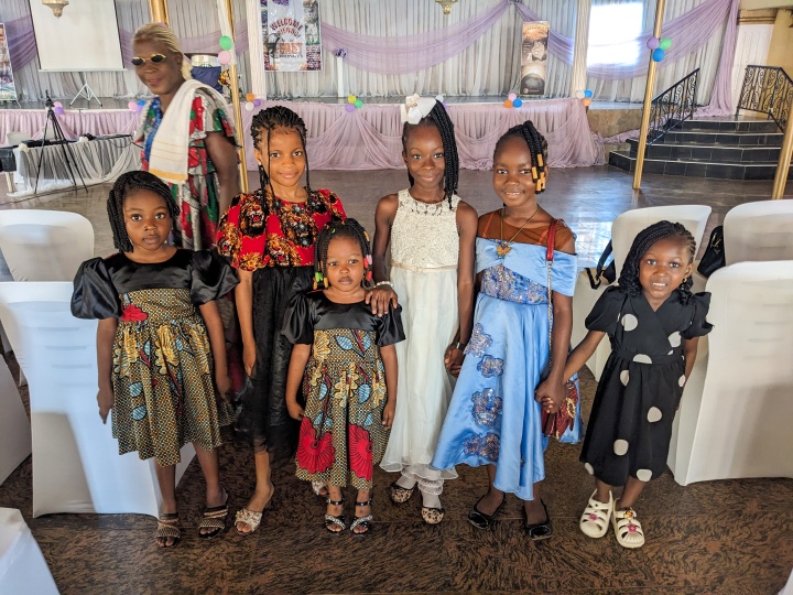 a group of girls wearing fancy dresses standing indoors with a lady walking the background