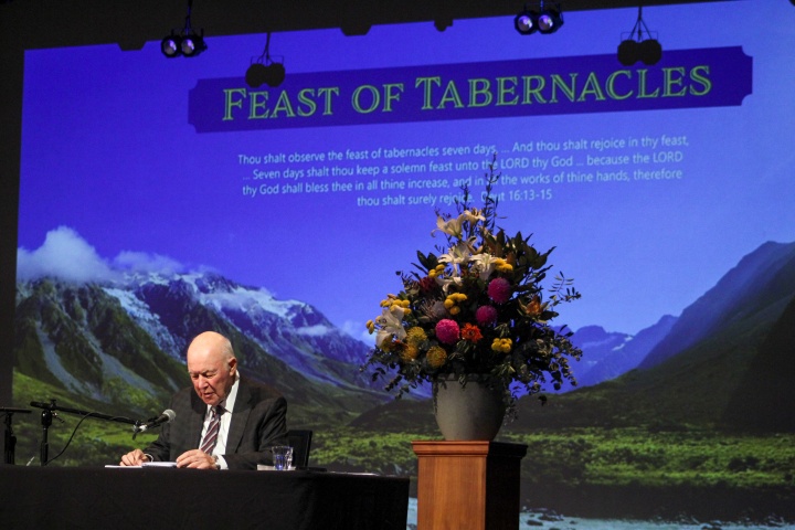 a man standing at the podium with a Feast of Tabernacles powerpoint presentation projected behind him