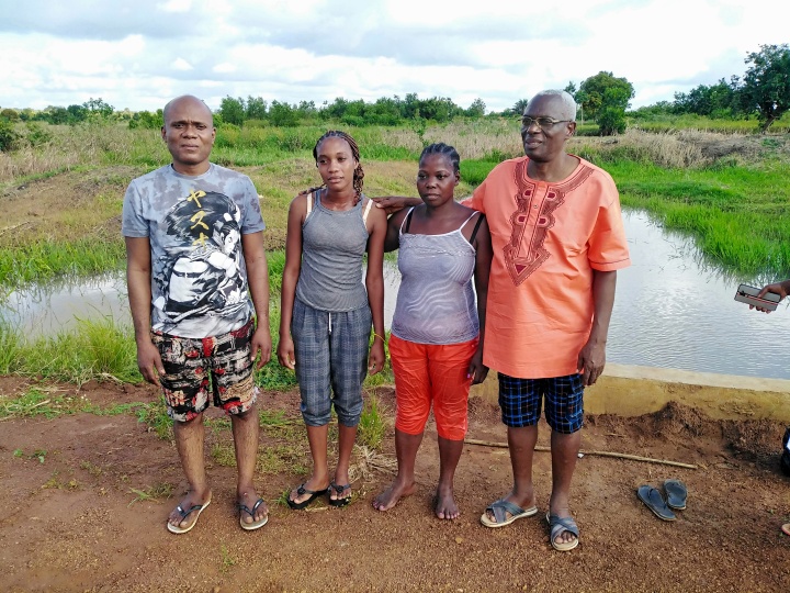 four people standing outside with a small pond in the background