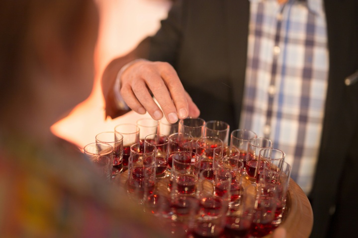 a man's hand reaching for a shot glass with red wine on a tray of similar glasses