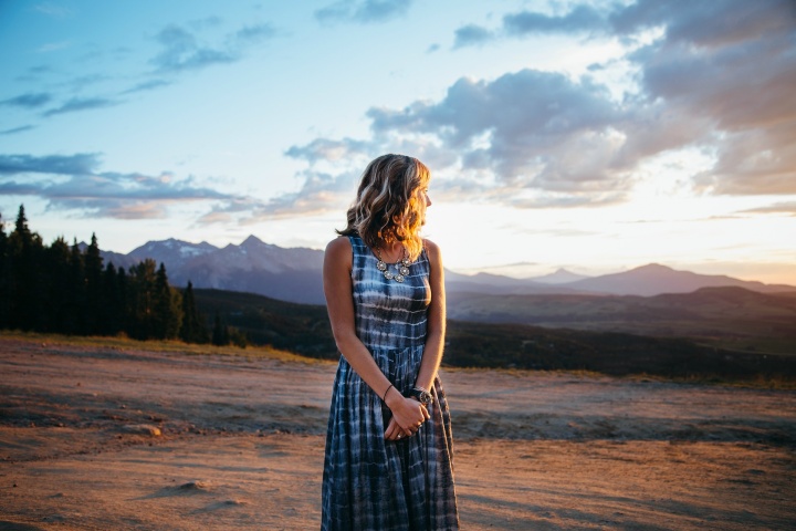 a woman wearing a long dress looking towards the sunset and she stands outside in a field