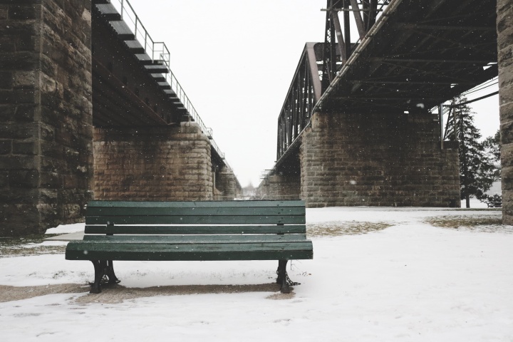 A park bench surrounded by snow and underneath a bridge.