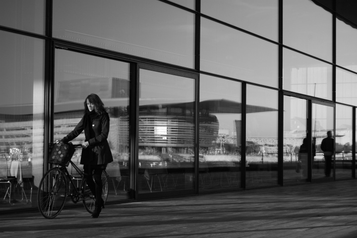 A young woman pushing a bike while walking by a office building with mirrored windows.