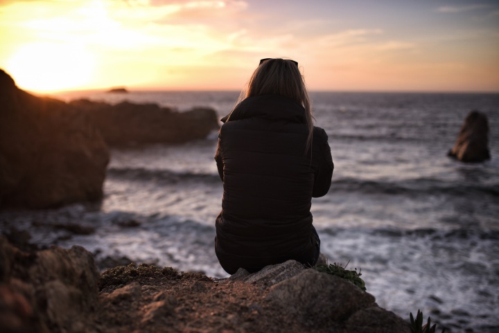 A young woman sitting on an rock looking at the sun setting over the ocean.